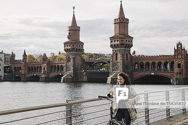 Lächelnde Frau mit Fahrrad und Smartphone in der Stadt an der Oberbaumbrücke  Berlin  Deutschland