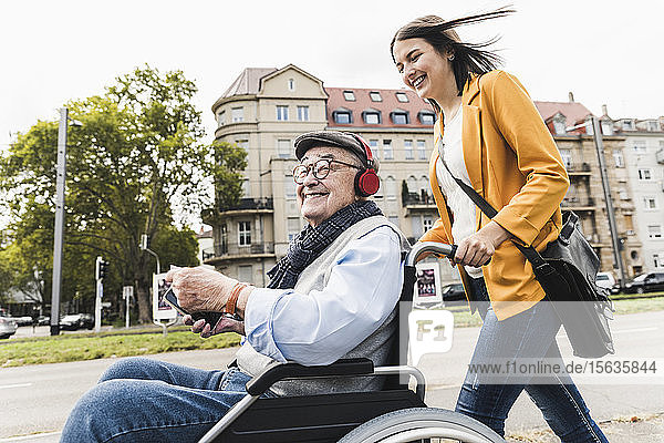Lachende junge Frau schiebt glücklichen älteren Mann mit Kopfhörern und Smartphone im Rollstuhl