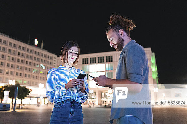 Happy couple in the city at night checking on their phones  Berlin  Germany