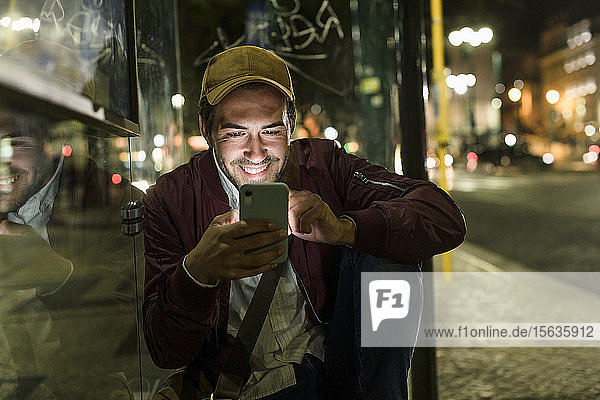 Porträt eines lächelnden jungen Mannes der nachts an einer Bushaltestelle sitzt und sein Handy benutzt Lissabon Portugal