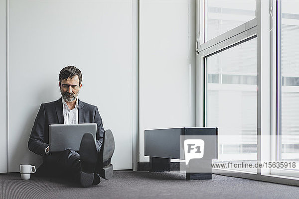 Reifer Geschäftsmann im Büro  der auf dem Boden sitzt und einen Laptop benutzt