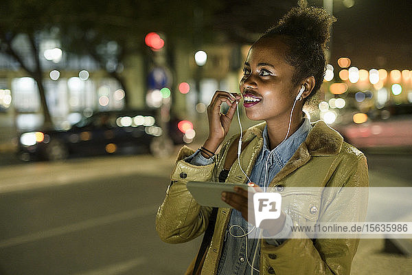 Porträt einer lächelnden jungen Frau mit Kopfhörer und Smartphone in der Stadt bei Nacht  Lissabon  Portugal