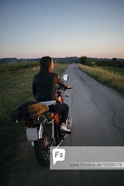 Rückenansicht einer jungen Frau  die bei Sonnenuntergang auf einem Motorrad sitzt und auf die Aussicht schaut  Toskana  Italien