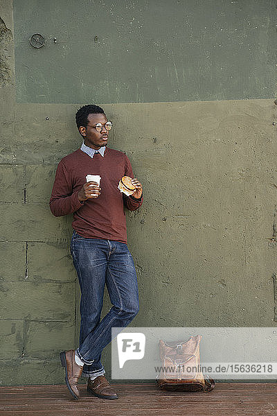 Junger Mann mit einem Chesseburger und Kaffee zum Mitnehmen  an der Wand gelehnt