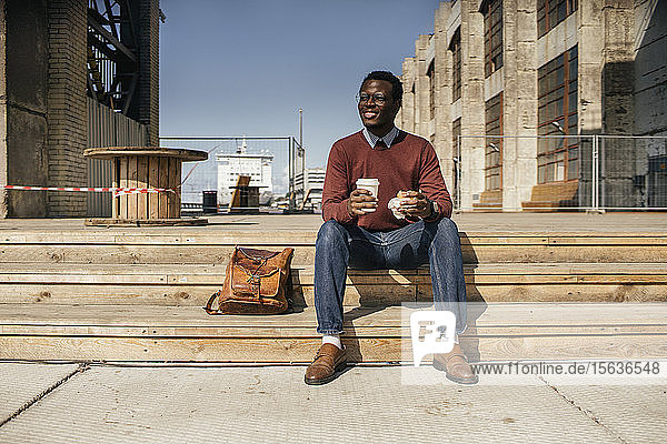 Junger Mann sitzt auf einer Treppe in der Stadt  isst Hamburger  trinkt Kaffee