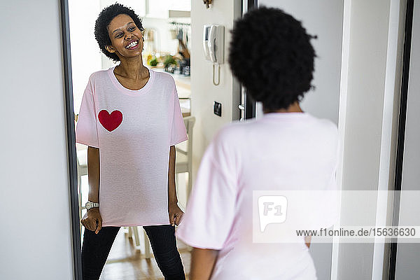 Glückliche junge Frau  die ein T-Shirt mit Herzform trägt und in den Spiegel schaut