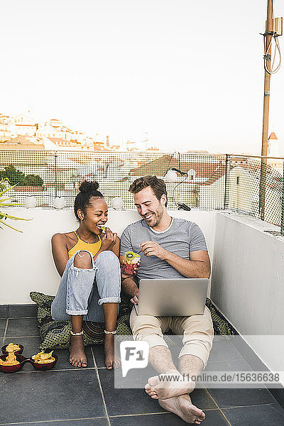 Happy young couple with laptop sitting on rooftop in the evening having a snack