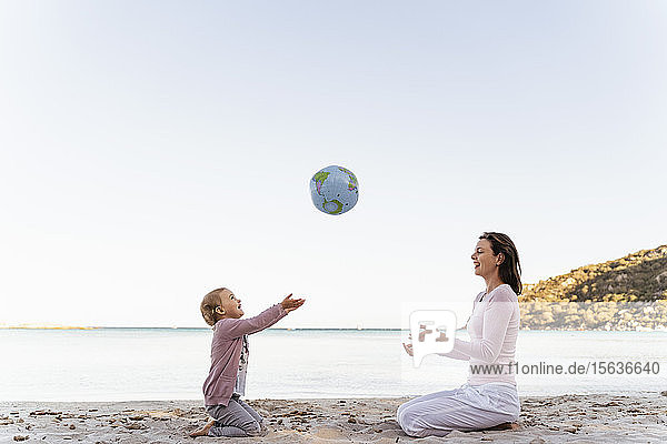 Glückliches kleines Mädchen sitzt mit seiner Mutter am Strand und spielt mit dem Earth Beachball