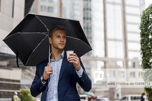 Junger Geschäftsmann mit Regenschirm trinkt an einem regnerischen Tag in Bangkok einen Kaffee