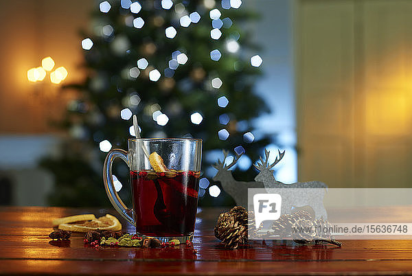 Nahaufnahme von Glühwein mit Essen und Dekorationen auf Holztisch vor beleuchtetem Weihnachtsbaum zu Hause