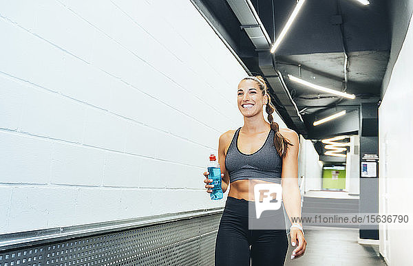 Lächelnde Frau in guter Verfassung geht während des Trainings mit Wasserflasche durch den Korridor