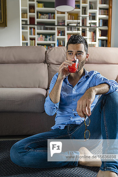 Portrait of young man sitting on the floor at home drinking glass of juice