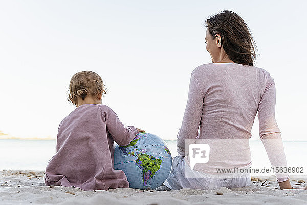 Rückenansicht eines kleinen Mädchens mit einem Strandball der Erde  das mit seiner Mutter am Meeresufer sitzt