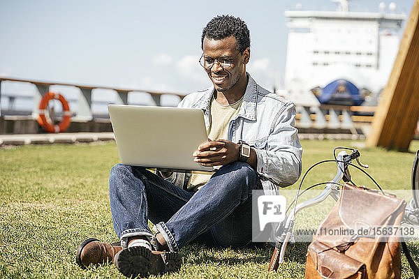 Junger Mann sitzt auf Rasen und benutzt Laptop