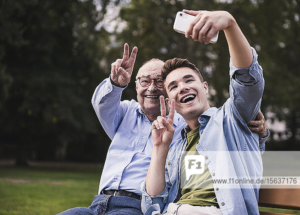 Älterer Mann und Enkel sitzen zusammen auf einer Parkbank und nehmen Selfie mit Smartphone