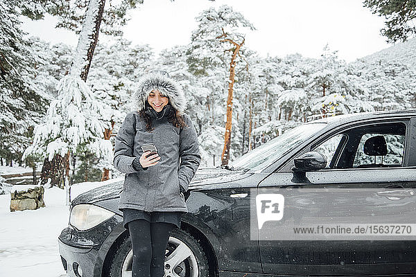 Porträt einer lächelnden jungen Frau  die sich im Winterwald an ein geparktes Auto lehnt und auf ihr Handy schaut
