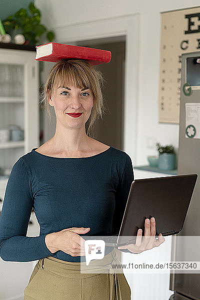 Porträt einer lächelnden Frau  die mit Laptop in der Küche steht und ein Buch auf ihrem Kopf balanciert