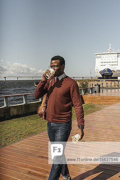 Junger Mann mit Kaffeetasse und Hamburger beim Spaziergang am Hafen