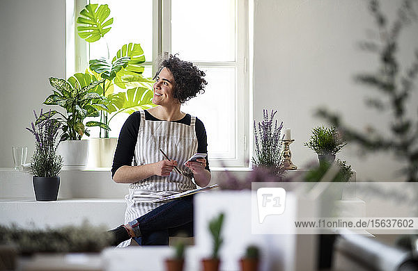 Lächelnde junge Frau mit Klemmbrett und Handy in einem kleinen Laden mit Pflanzen