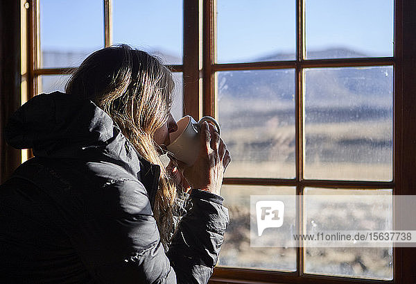 Frau trinkt eine Tasse Kaffee  während sie durch das Fenster die Berge bewundert
