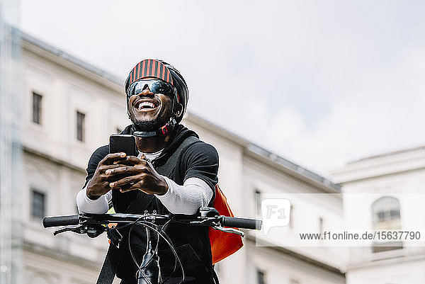 Glücklicher stilvoller junger Mann mit Fahrrad  Smartphone und Kuriertasche in der Stadt