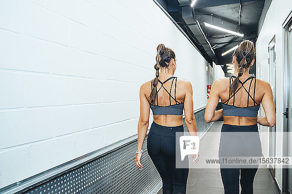 Rückansicht gut erhaltener weiblicher Zwillinge  die während des Trainings in einer Turnhalle durch den Korridor gehen