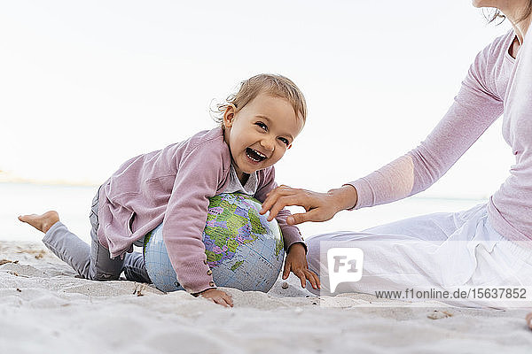 Porträt eines fröhlichen kleinen Mädchens  das mit dem Earth Beachball am Strand spielt