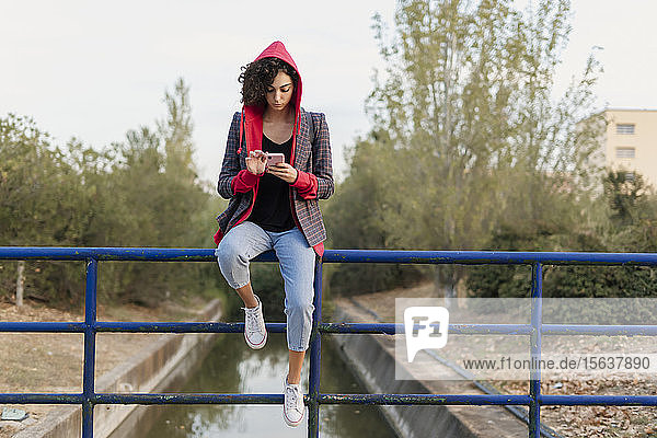 Junge Frau sitzt auf einem Geländer und benutzt ein Mobiltelefon