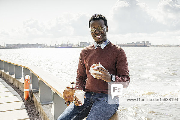 Young man sitting on railing at the sea  eating hamburger  drinking coffee