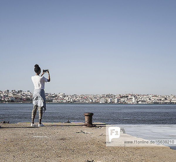 Junge Frau steht am Pier am Wasser und macht ein Handyfoto  Lissabon  Portugal