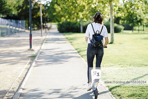 Rückenansicht einer Frau mit Rucksack auf einem Elektroroller im Stadtpark