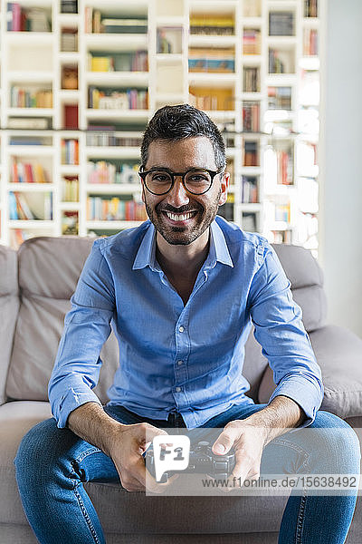 Porträt eines lachenden jungen Mannes  der zu Hause auf der Couch sitzt und ein Videospiel spielt