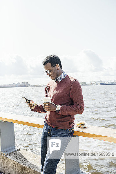 Junger Mann steht am Meer  hält Kaffee in der Hand und benutzt ein Smartphone