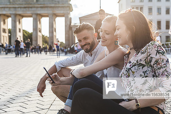 Drei glückliche Freunde sitzen in der Nähe des Brandenburger Tors und schauen auf ihr Handy  Berlin  Deutschland