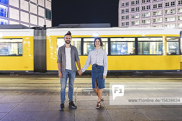 Glückliches Paar hält sich an den Händen und schaut in die Kamera mit der Straßenbahn im Hintergrund  Berlin  Deutschland