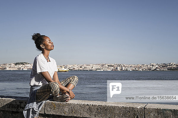 Young woman sitting on a wall at the waterfront with closed eyes  Lisbon  Portugal