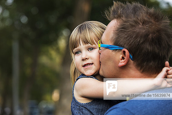 Portrait of a father cuddling with his young daughter and kissing her on the cheek; Edmonton  Alberta  Canada