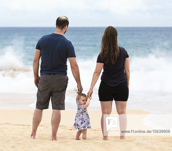 Mother and father hold the hands of their baby daughter as they stand looking out at the ocean; Maui  Hawaii  United States of America