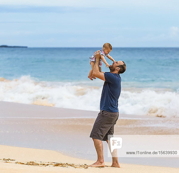 Father holds his baby daughter in the air as he stands on the beach next to the ocean; Maui  Hawaii  United States of America