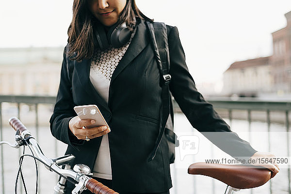 Mittelsektion einer Geschäftsfrau  die ein Mobiltelefon benutzt  während sie mit dem Fahrrad auf einer Brücke in der Stadt steht