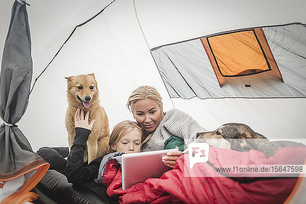Mother and daughter watching digital tablet while lying with pets in tent at camping site