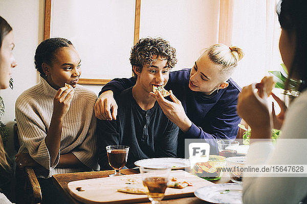 Teenager feeding pizza to friend while females looking at them in living room