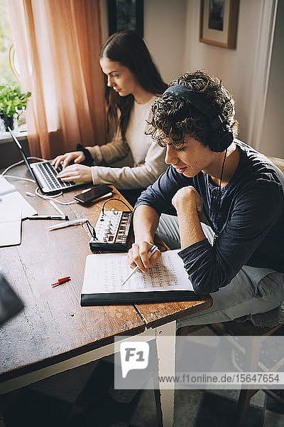 Hochwinkelansicht eines Teenagers  der mit Kopfhörern Musik hört  während ein Freund einen Laptop am Tisch im Haus benutzt