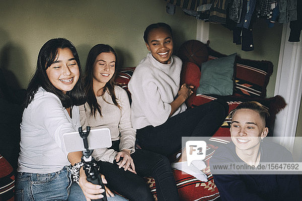 Lächelndes Teenager-Mädchen  das sich mit Freunden auf einem Einbeinstativ selbstständig macht  während es sich zu Hause vergnügt