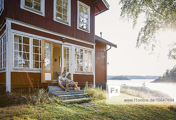 Full length of couple sitting at log cabin entrance by lake during summer