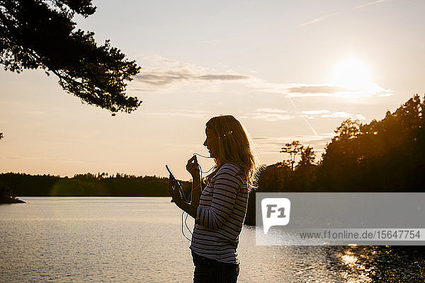 Mittelgroße erwachsene Frau  die während des Sonnenuntergangs am See mit Kopfhörern über Handy telefoniert