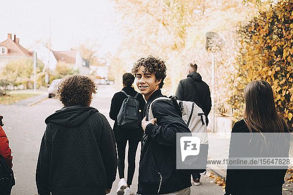 Portrait of smiling teenage boy carrying backpack while walking with friends on street in city