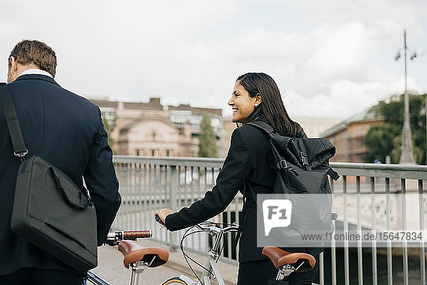 Smiling businesswoman looking at businessman while walking with bicycles on bridge in city