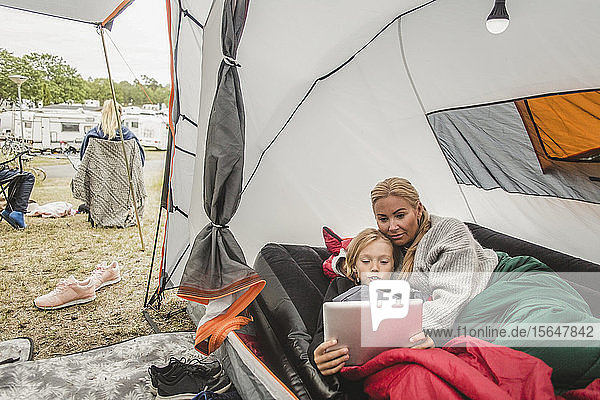 Mädchen sieht sich ein digitales Tablet an  während sie mit der Mutter im Zelt auf dem Campingplatz liegt