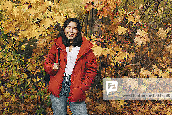 Porträt eines lächelnden Teenager-Mädchens  das mit Rucksack vor herbstlichen Bäumen steht